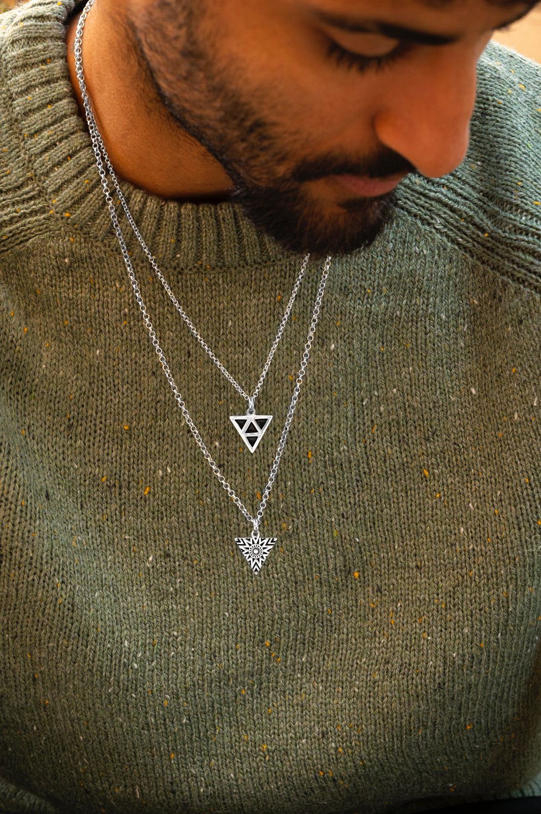 Man wearing two silver necklaces with triangle pendants on a green sweater.